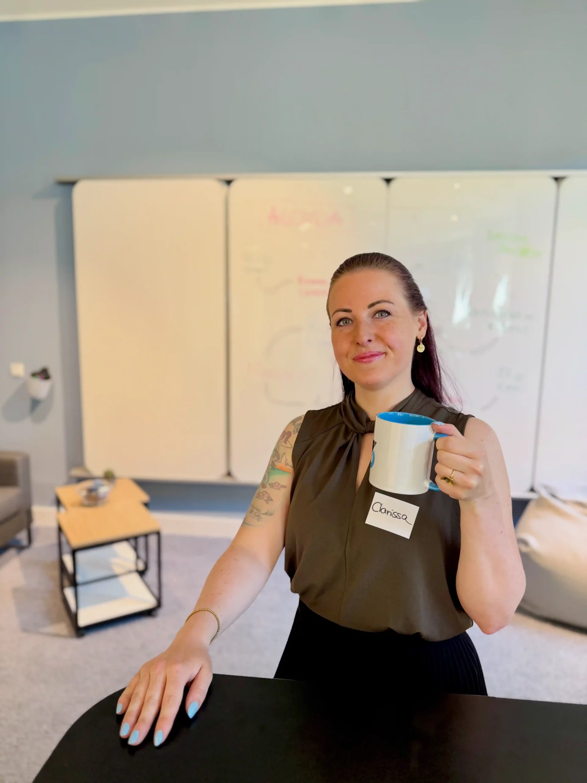 Frau Clarissa Muenchow mit langen, dunklen Haaren hält eine Tasse in der Hand. Sie trägt ein ärmelloses, dunkelbraunes Oberteil und hat einen Namensschild mit 'Clarissa' an ihrem Oberteil. Im Hintergrund sind ein Tisch, ein Stuhl und eine beschreibbare Wandtafel zu sehen.