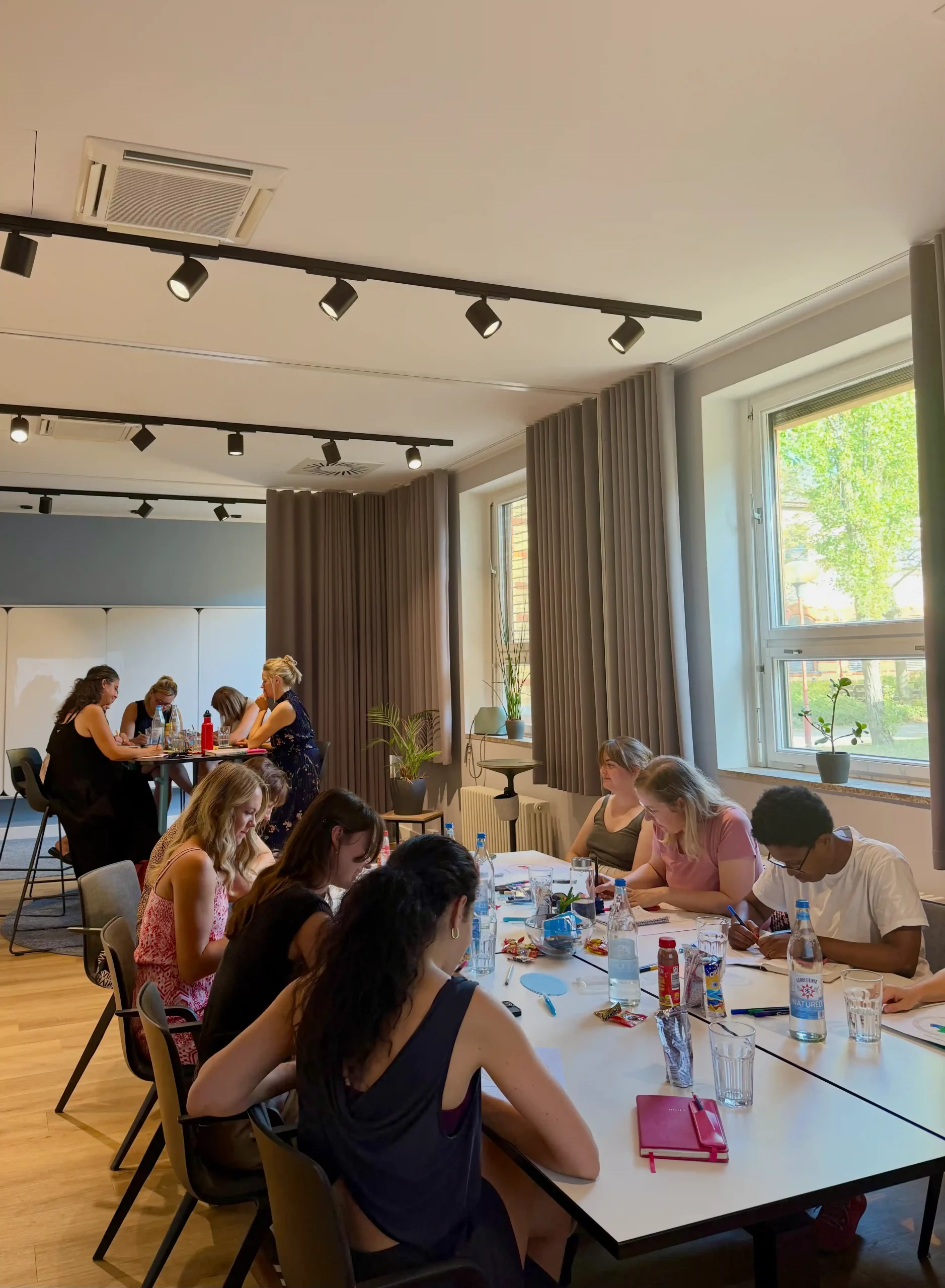Gruppensitzung in einem Workshop mit mehreren Teilnehmerinnen, die an einem Tisch sitzen und arbeiten. Im Hintergrund stehen weitere Personen, die an einem Flipchart arbeiten. Fenster mit Tageslicht.